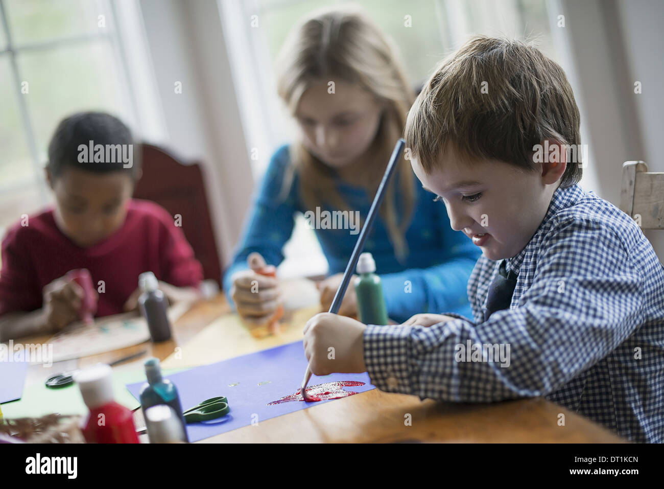 I bambini in una casa famiglia di tre bambini seduti a un tavolo utilizzando colla e vernice per creare decorazioni Foto Stock