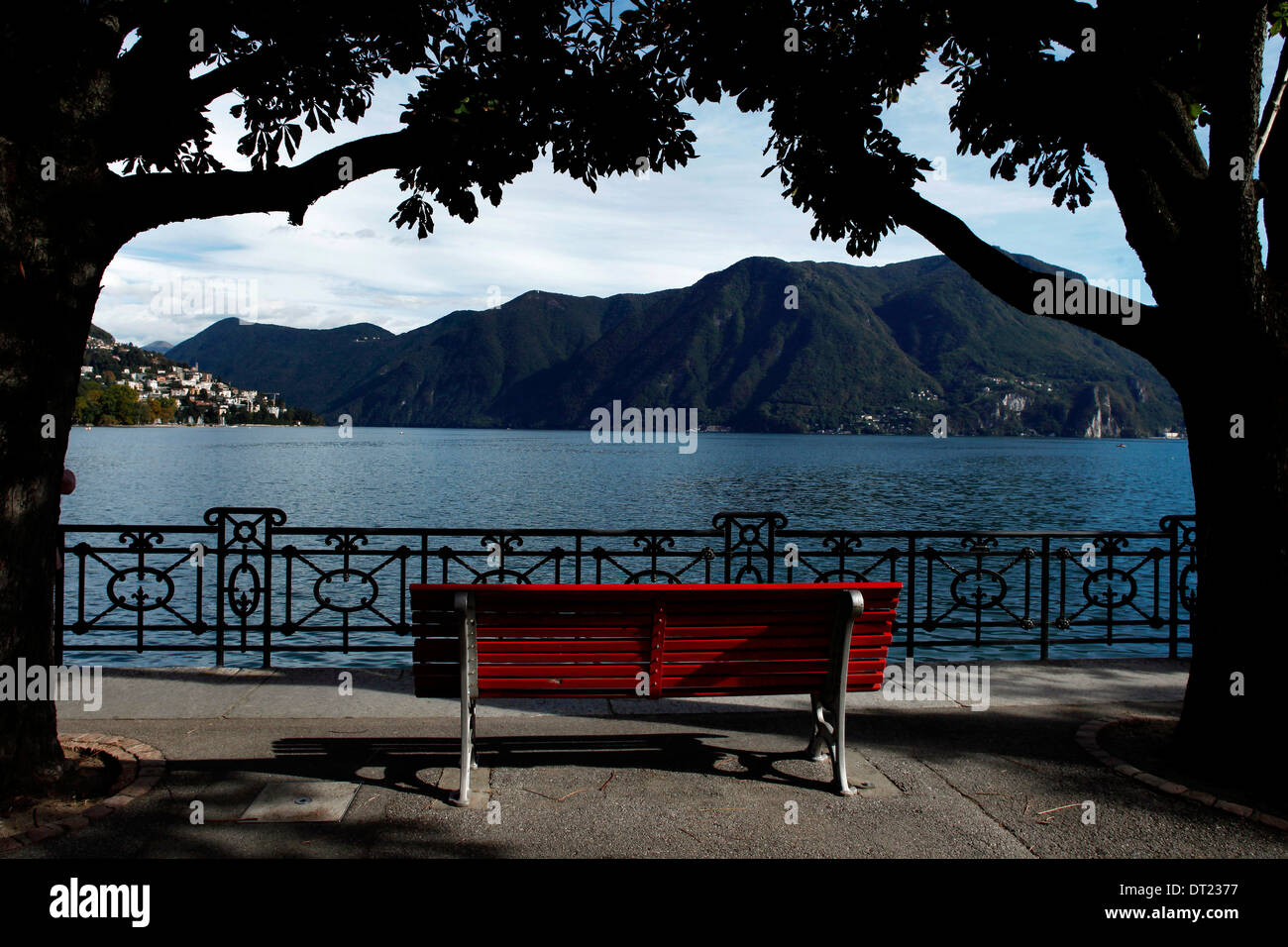 Un banco di rosso tra due alberi di fronte lago di Lugano Foto Stock