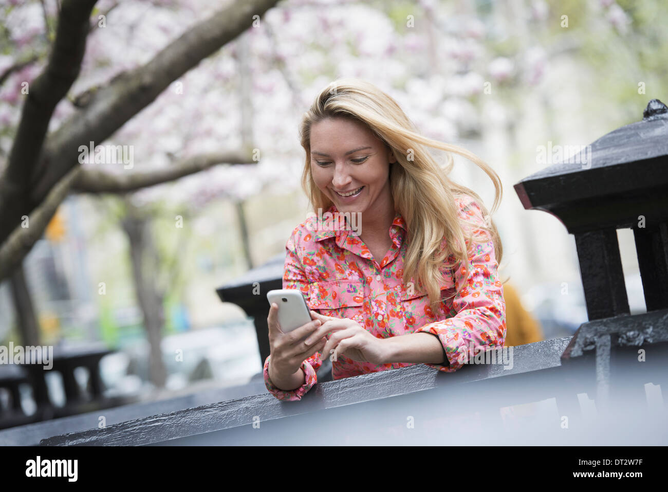 Vista su cityA giovane donna con capelli lunghi biondi seduti in un parco della città guardando il suo smart phone Foto Stock