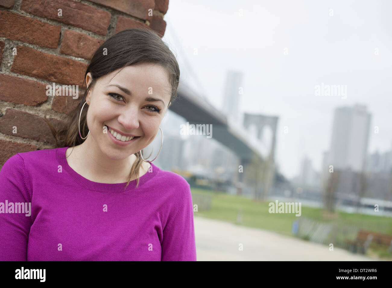 Ponte di Brooklyn attraversando la East River una donna seduta appoggiata contro un muro di mattoni controllando il suo smart phone Foto Stock