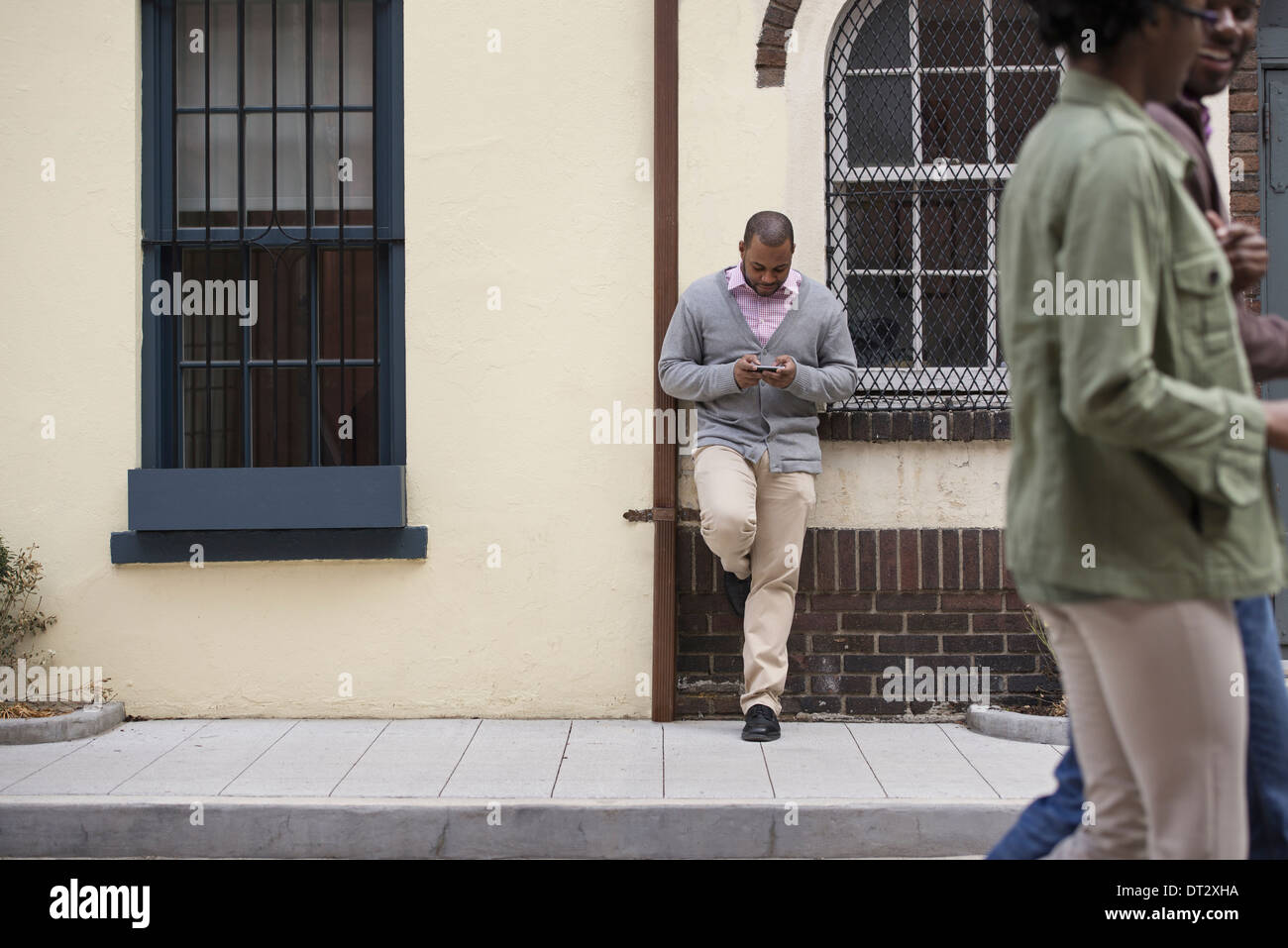 All'aperto nella città in primavera un giovane camminando sul marciapiede e un uomo appoggiato contro una parete controllando il suo telefono Foto Stock