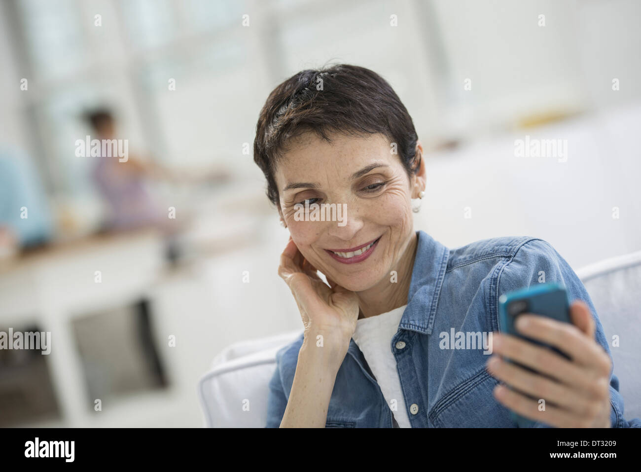 Professionisti in ufficio una luminosa e ariosa e luogo di lavoro di una donna matura in un blu denim shirt guardando un Blu smart phone Foto Stock