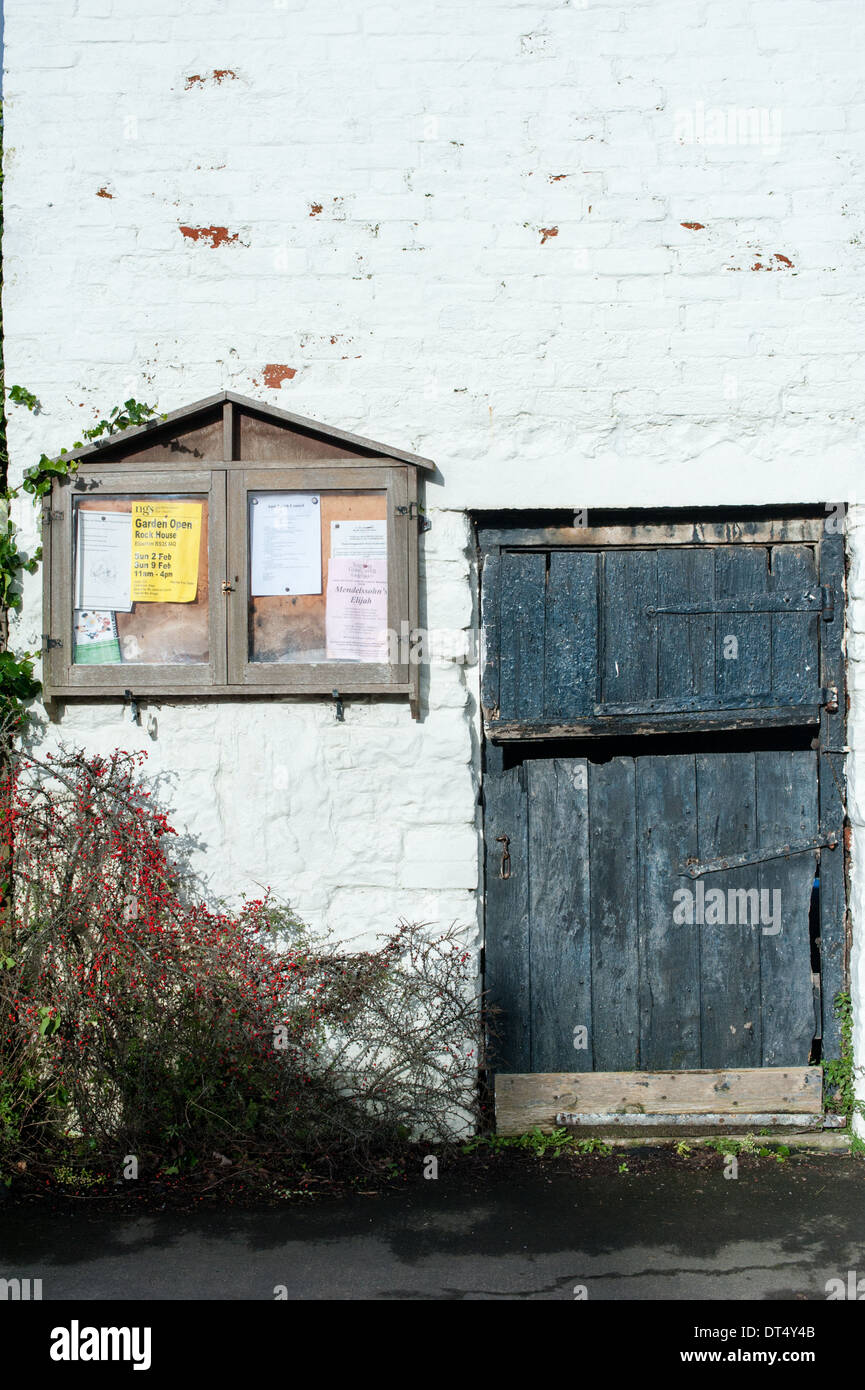 Nero antico stabile in legno porta accanto ad un villaggio bacheca su un dipinto di bianco muro di pietra nelle zone rurali di South Gloucestershire Foto Stock
