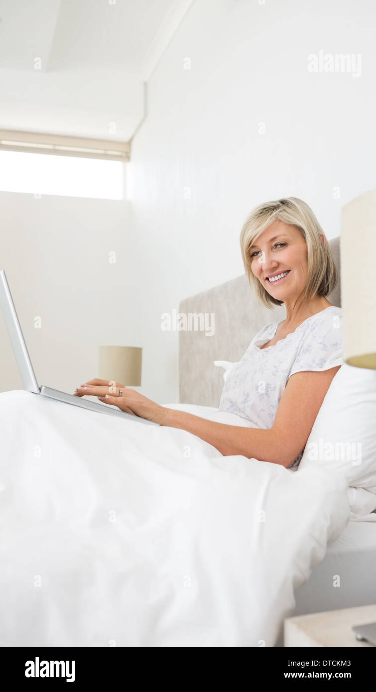 Sorridente donna matura utilizzando laptop a letto Foto Stock