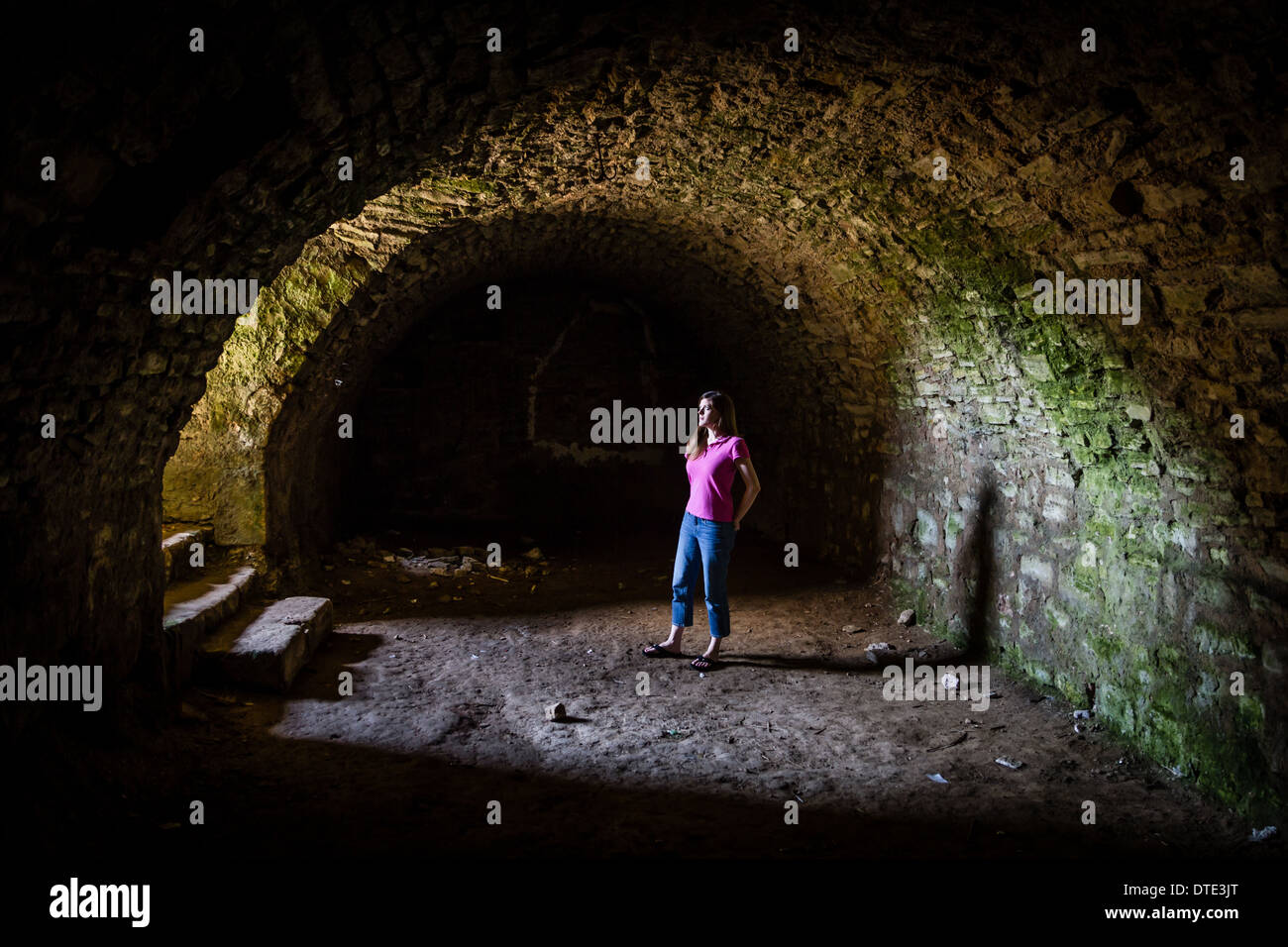 Ragazza giovane e carina sorge in un tunnel masoned nella cantina di un castello di monumenti medievali e guarda su per le scale in luce incidente. Foto Stock