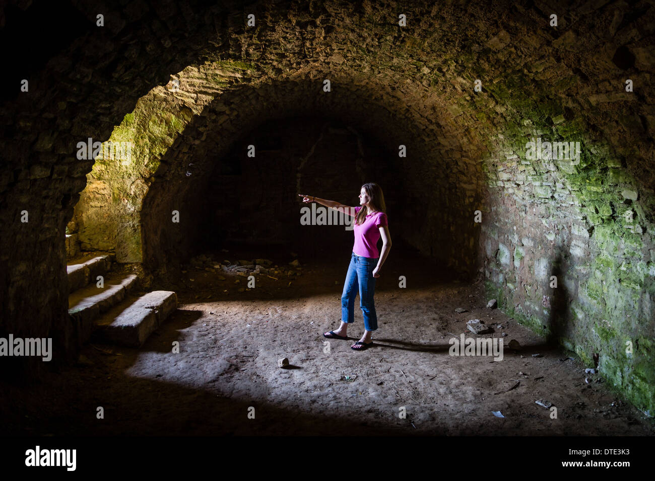 Ragazza giovane e carina sorge in un tunnel masoned nella cantina di un castello di monumenti medievali e punti su per le scale in luce incidente. Foto Stock