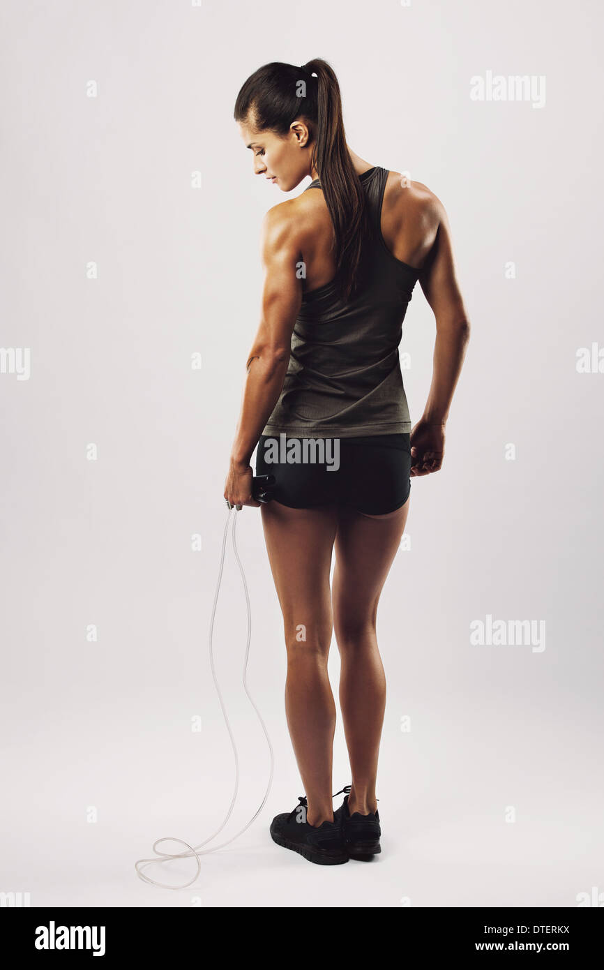 Montare e muscolare di donna con salto con la corda in piedi su sfondo grigio. Immagine della vista posteriore di bodybuilder femminile tenendo la corda. Foto Stock