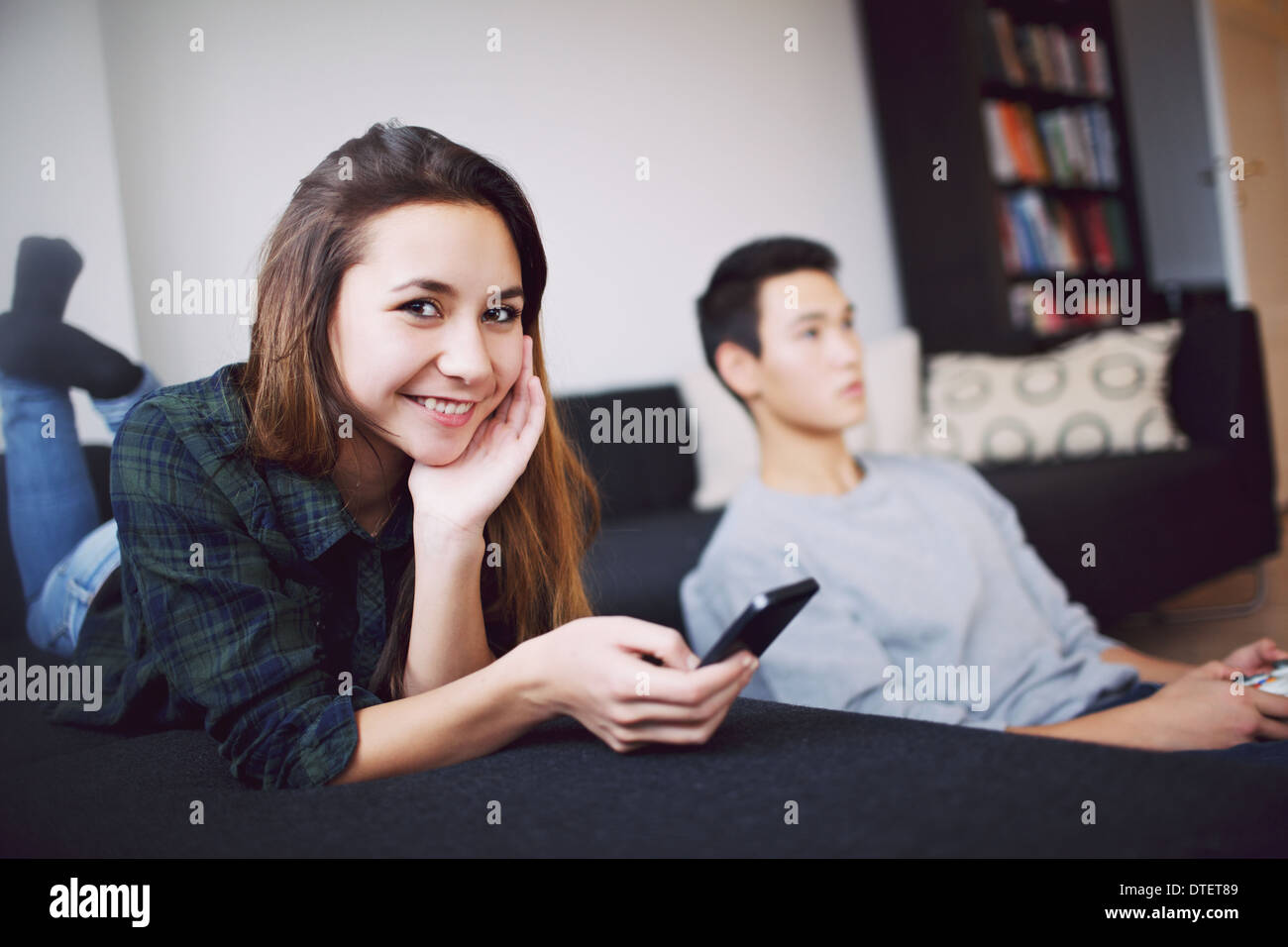 Attraente ragazza adolescente sdraiato sul divano tenendo un telefono intelligente guardando la telecamera sorridendo. Giovane uomo seduto riproduzione in background. Foto Stock