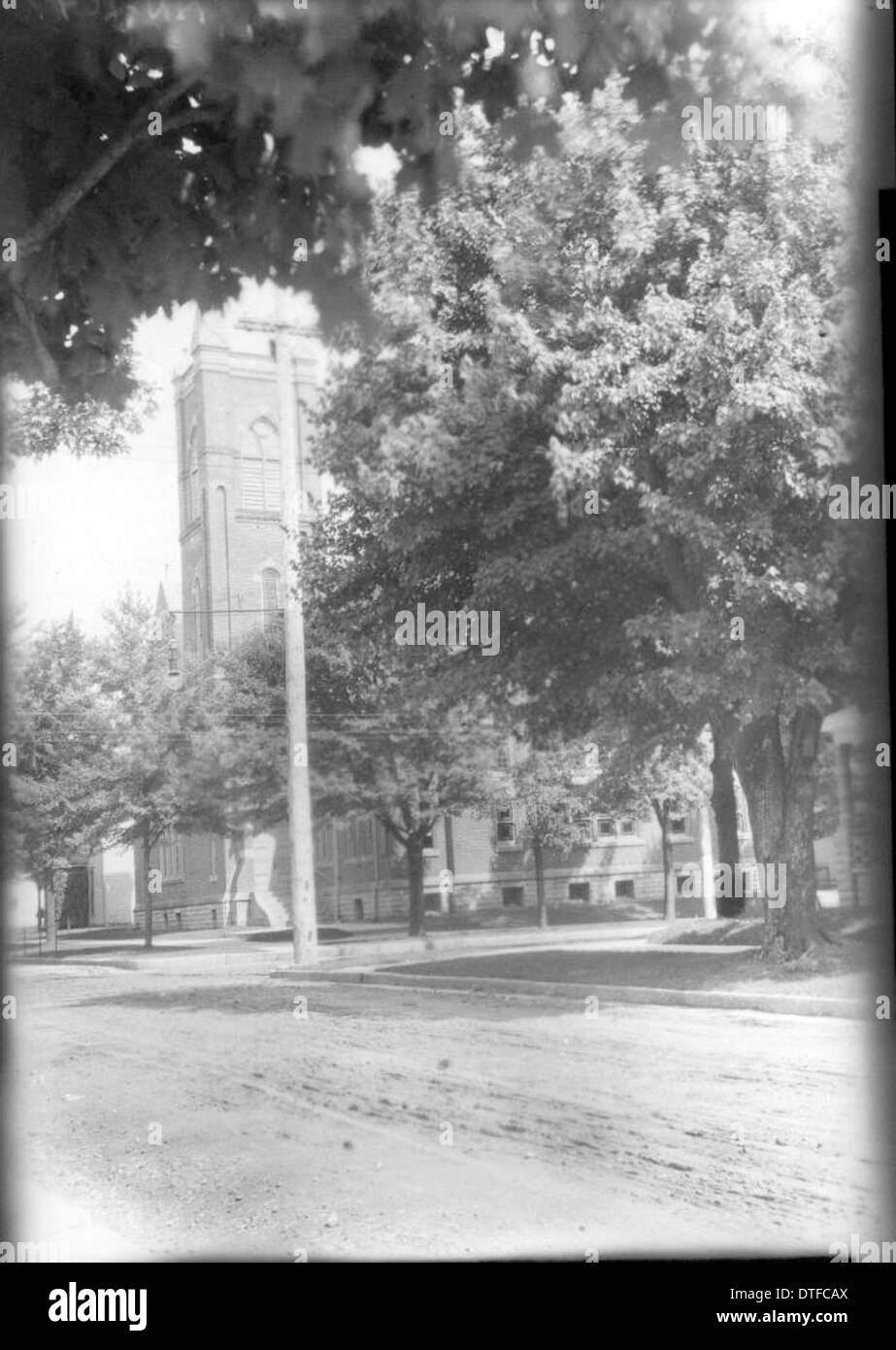 Una fotografia d'epoca con una vista parzialmente oscurata di una chiesa attraverso gli alberi. Questa cartolina cattura una scena tranquilla e basata sulla natura, con il campanile della chiesa che emerge sottilmente da dietro il fogliame, mostrando tecniche fotografiche e caratteristiche architettoniche dell'inizio del XX secolo. Foto Stock