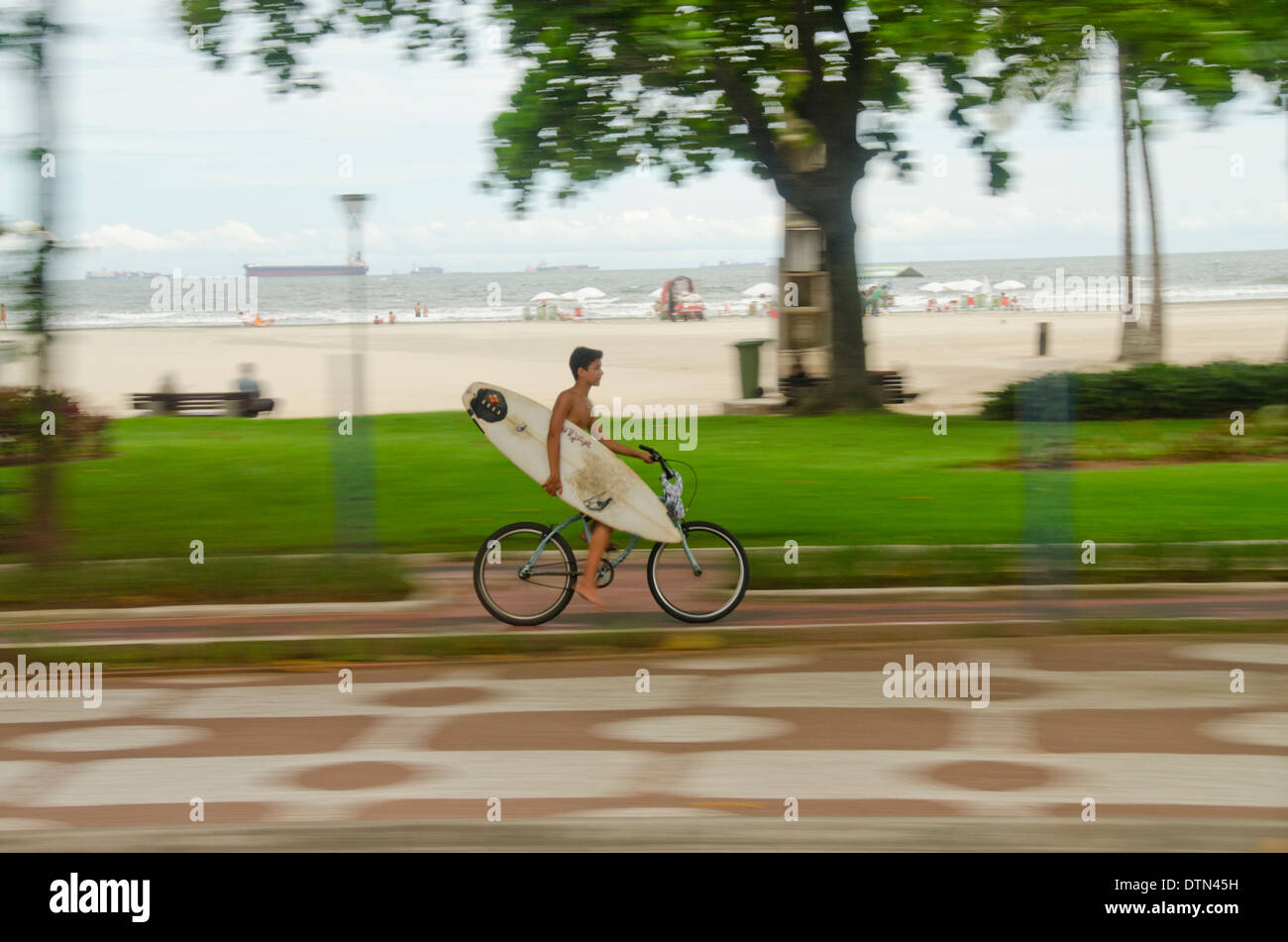 Il Brasile, Sao Paulo, Santos. Giovane ragazzo in bici che porta tavola da surf lungo il marciapiede sul lungomare. Foto Stock