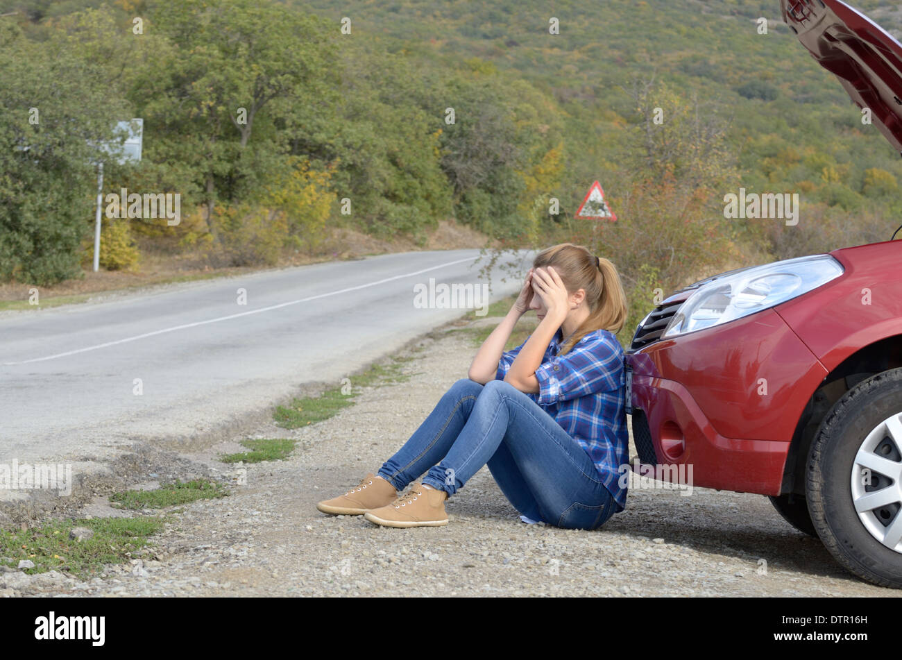 Giovane donna vicino a Broken auto necessita di assistenza seduto sotto il cofano aperto Foto Stock