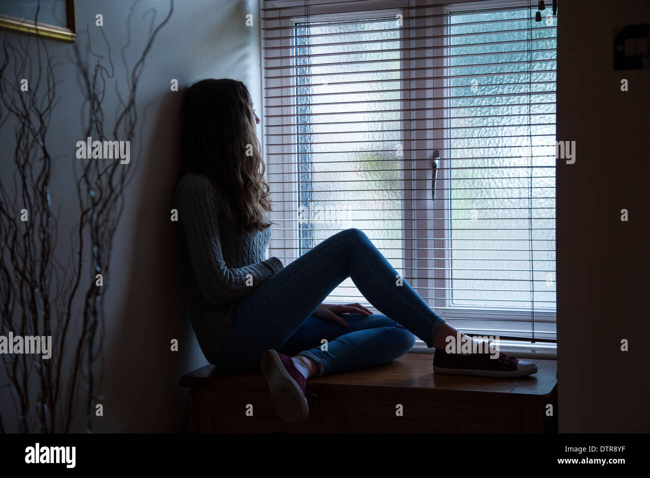 Giovane donna con capelli lunghi che indossa jeans, triste e guardando verso una finestra, seduti al buio a casa. Punto di vista laterale. Foto Stock