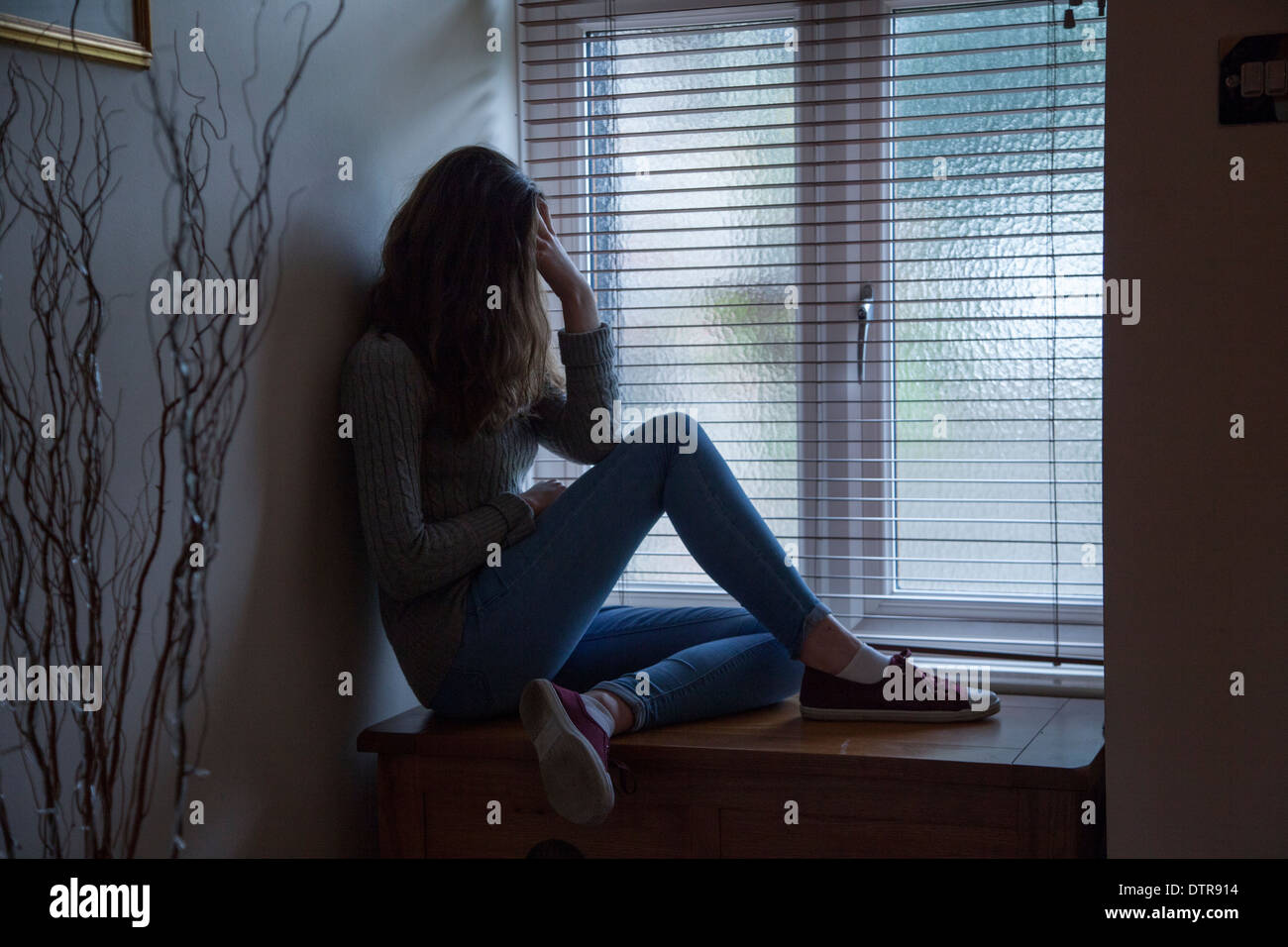 Giovane donna con capelli lunghi che indossa jeans, mano sulla testa nella disperazione, seduti nelle tenebre da una finestra a casa. Punto di vista laterale. Foto Stock