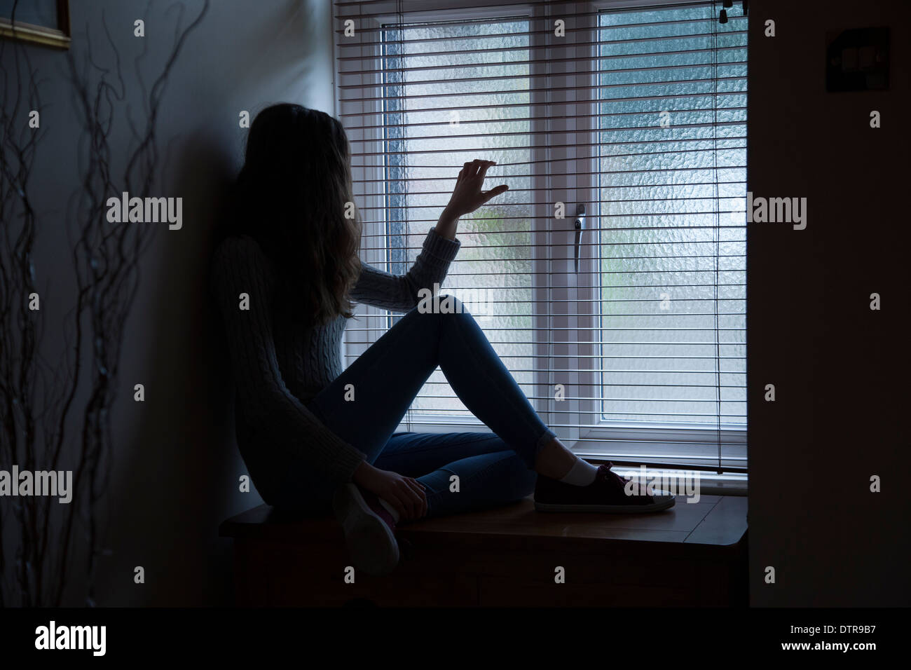 Irriconoscibile lonely ragazza con i capelli lunghi che indossa jeans di guardare attraverso una finestra cieca, seduti nelle tenebre. Foto Stock