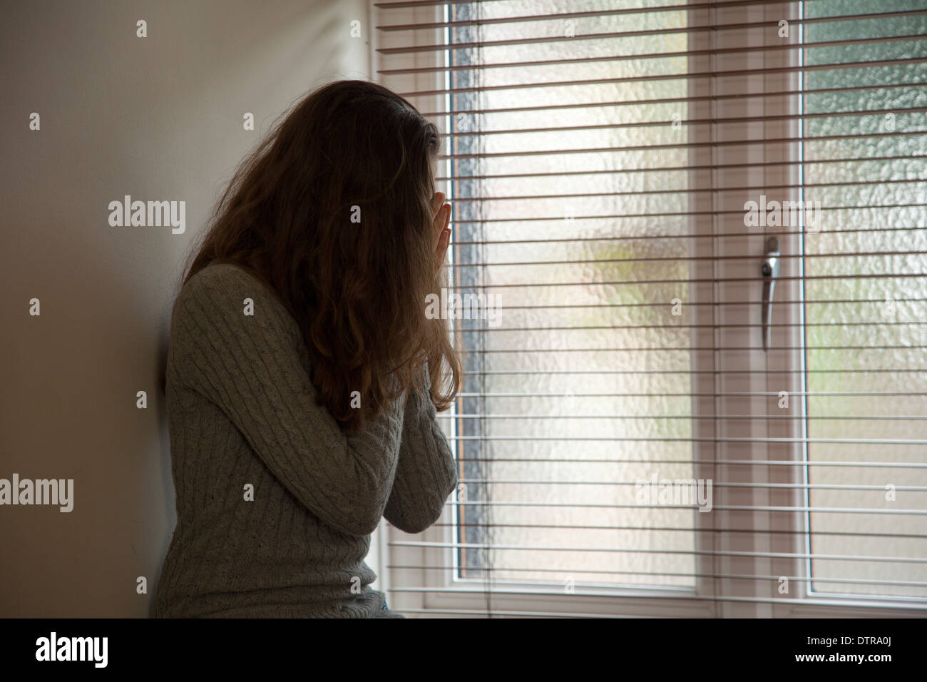 La ragazza con i capelli lunghi che indossa jeans, testa in mani, seduto da solo da una finestra di casa. Punto di vista laterale. Foto Stock