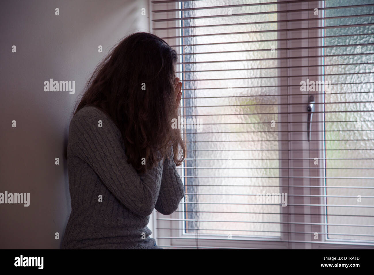 La ragazza con i capelli lunghi che indossa jeans, testa in mani, seduto da solo da una finestra di casa. Punto di vista laterale. Foto Stock