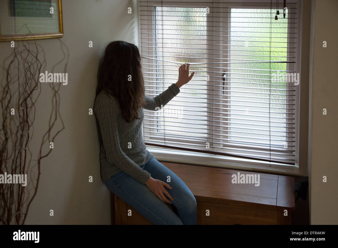 Irriconoscibile giovane donna con capelli lunghi che indossa jeans di guardare attraverso una finestra cieca. Foto Stock