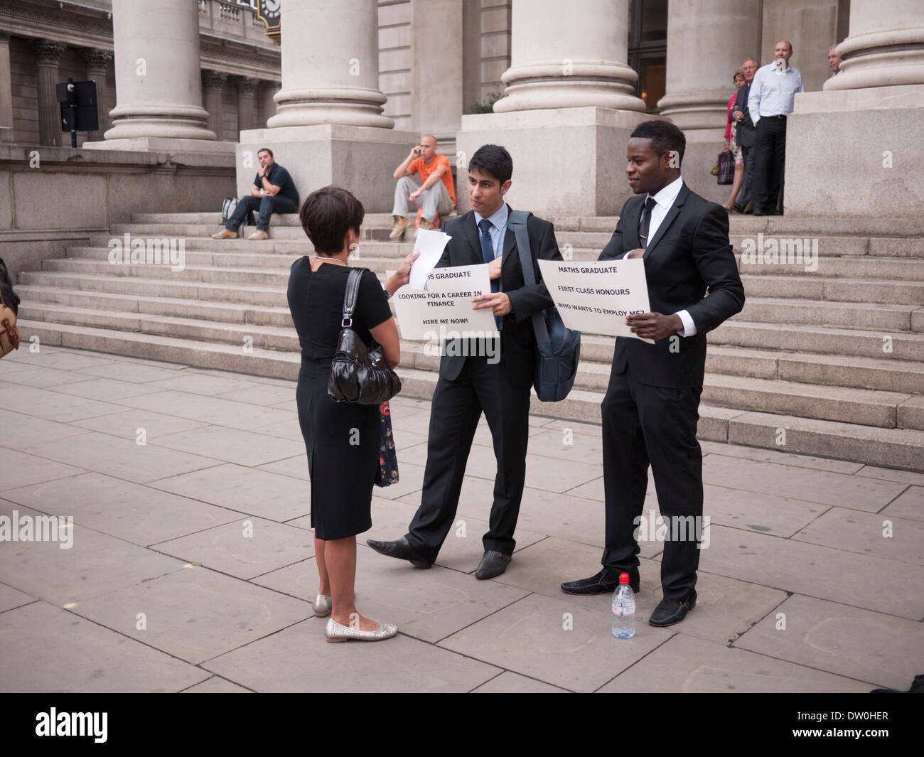 Laureati in matematica ben vestiti con cartelli che recano "Hire me Now" e "Who Wants to Employ me" che propongono lavori al di fuori del Royal Exchange nella City di Londra, Regno Unito. Neolaureati che cercano attivamente un impiego nel settore competitivo dei servizi finanziari e professionali. Foto Stock
