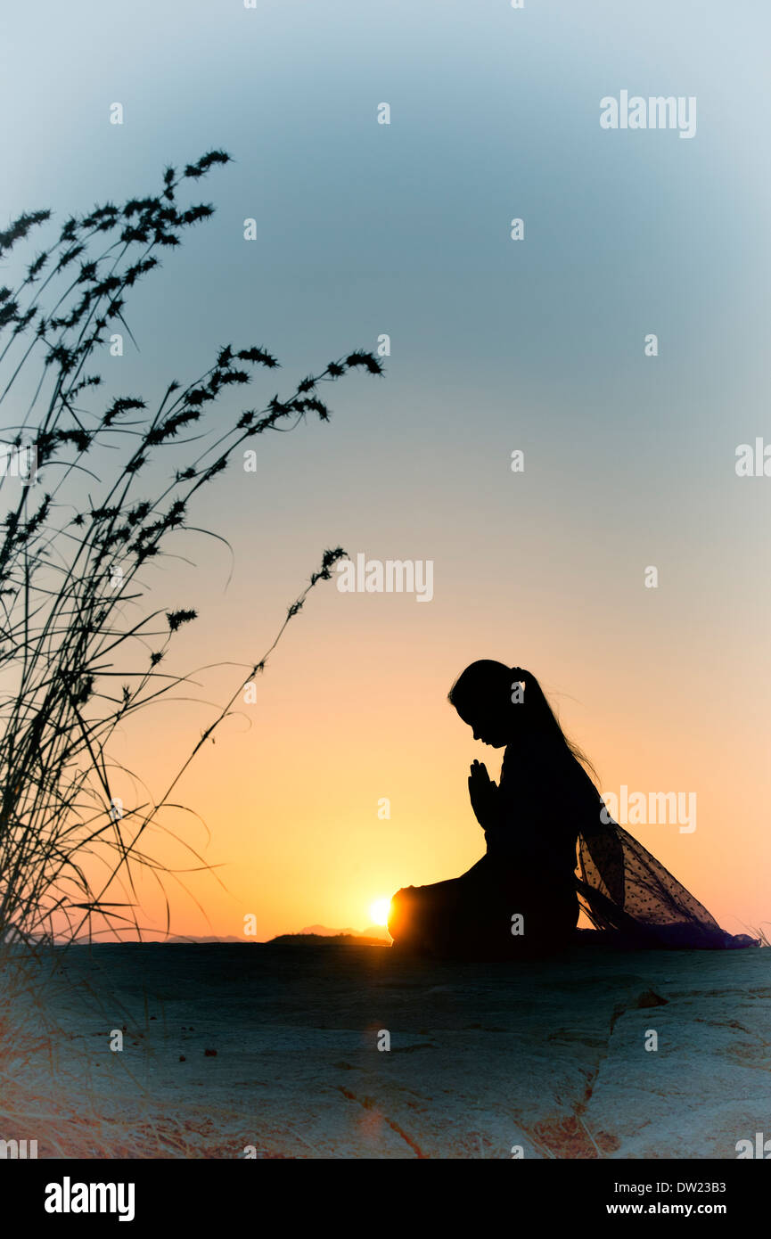 Indian ragazza adolescente pregando al tramonto nella campagna indiana. Silhouette. Con applicato il filtro nostalgico Foto Stock