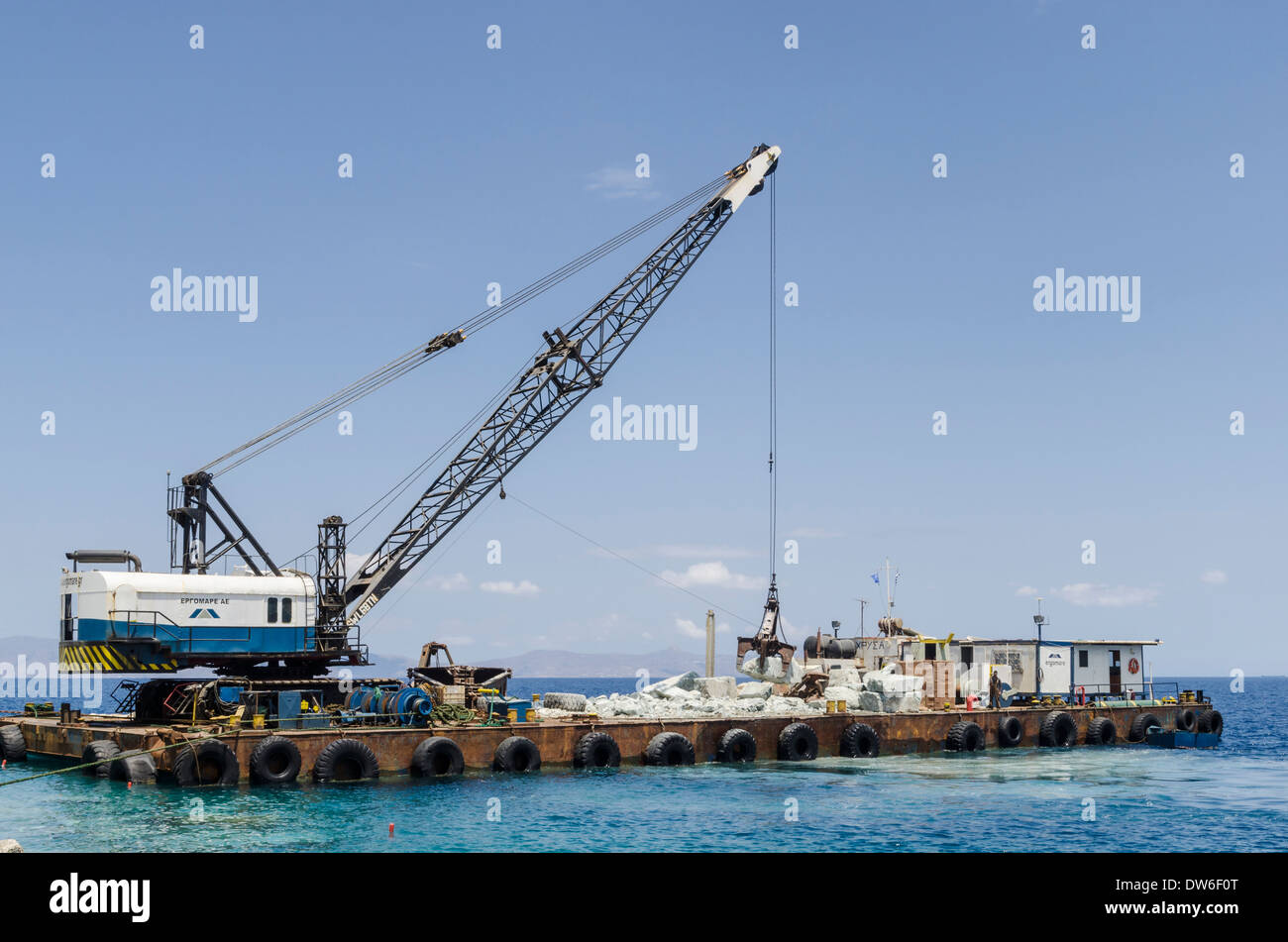Barge con gru di sollevamento di rocce per una parete del mare lungo il porto esterno muro in Syros Town, Grecia Foto Stock