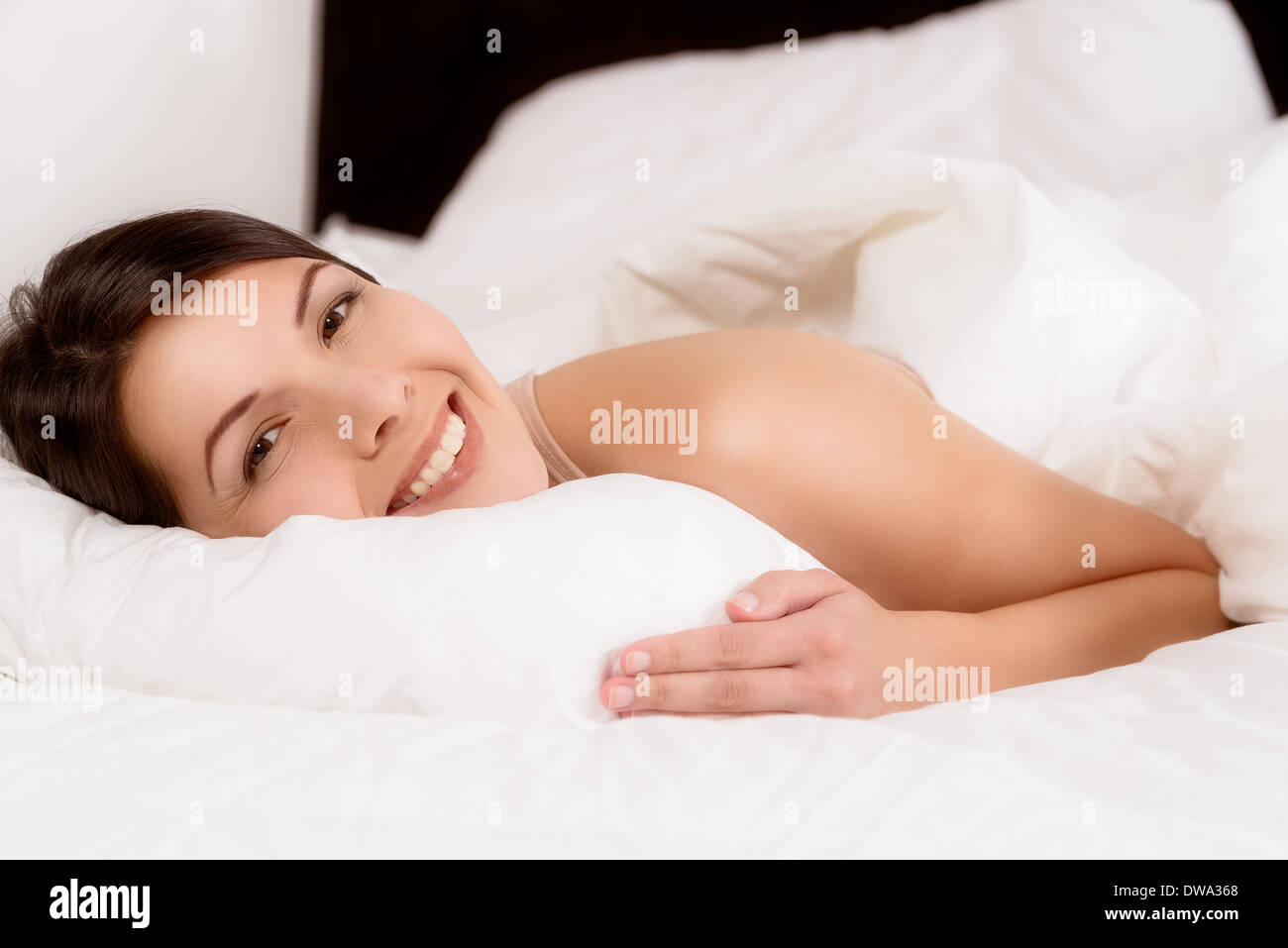 Sorridente ringiovanito sano giovane donna snuggling in basso tra i suoi cuscini nel letto dando alla telecamera un ampia trasmissione via IR di un sorriso di gioia Foto Stock