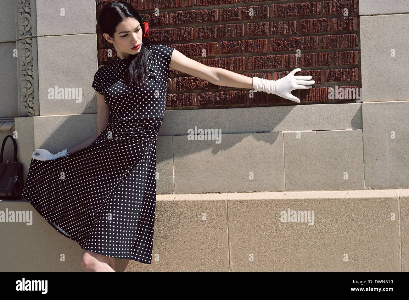 Un modello femminile magra contro una parete che indossa un vintage ispirato polka dot dress e guanto bianco tenendo alto il suo mantello Foto Stock