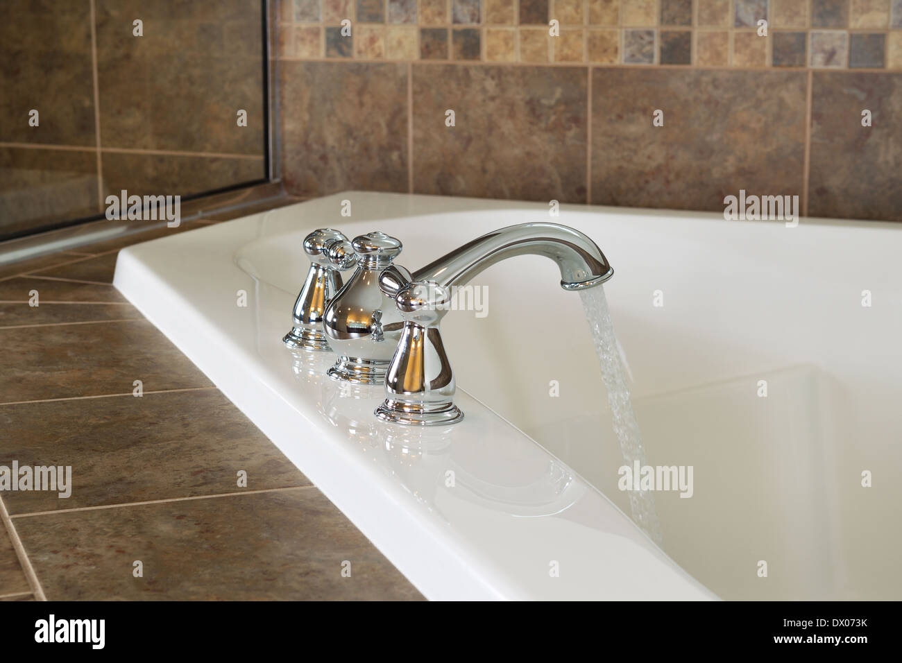 La foto in orizzontale del rubinetto cromato in funzione dell'acqua in vasca nella stanza da bagno principale con doccia parziale di vetro in background Foto Stock