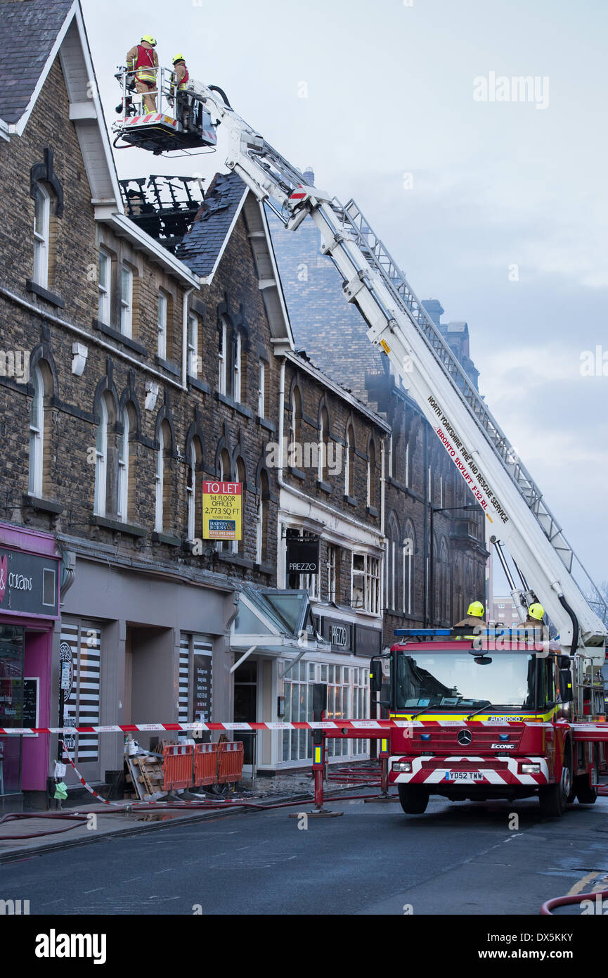 Coraggiosi vigili del fuoco alto della scala (2 al di sotto del motore) controllare i danni di un incendio al tetto in centro storico edificio - Harrogate, North Yorkshire, Inghilterra, Regno Unito. Foto Stock