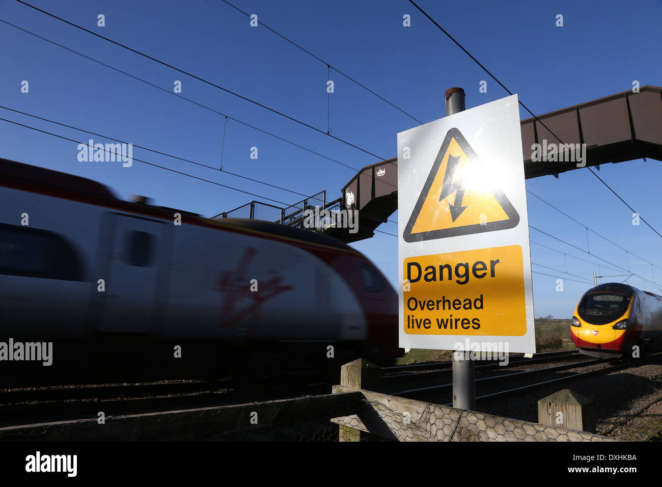 Pericolo cavi aerei sotto tensione segno accanto a una linea ferroviaria ponte pedonale come due treni Virign pass Foto Stock