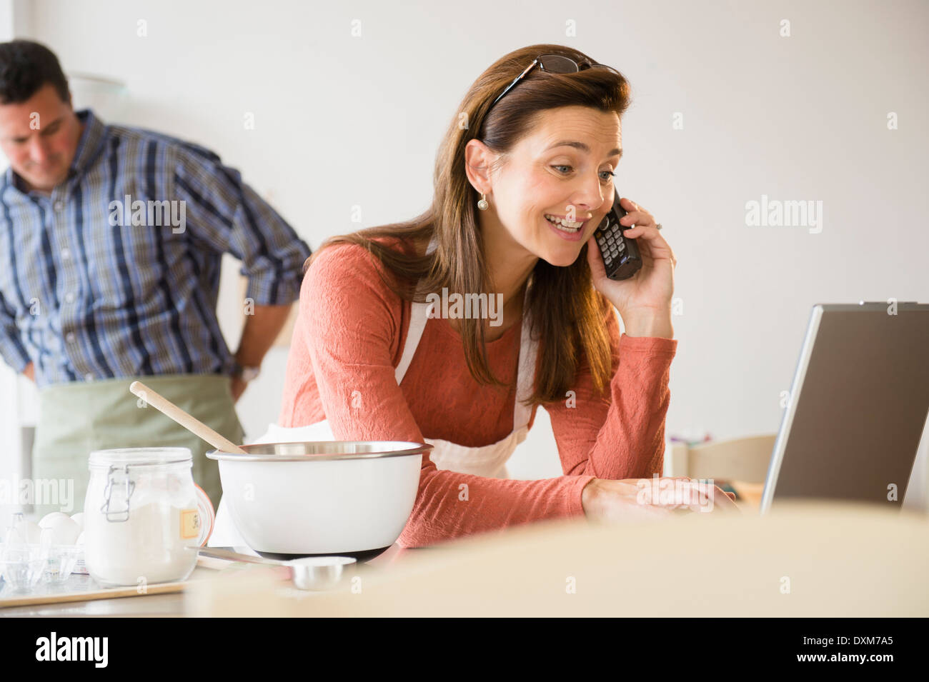 La donna caucasica la cottura, utilizzando laptop e parlando al telefono Foto Stock