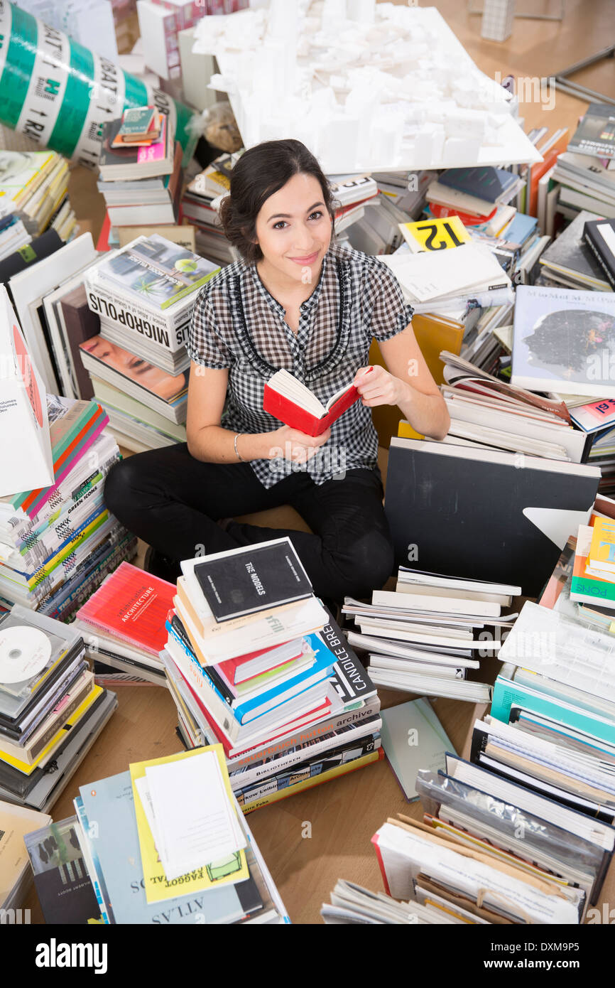 Sorridente giovane donna seduta al centro di pile di libri Foto Stock