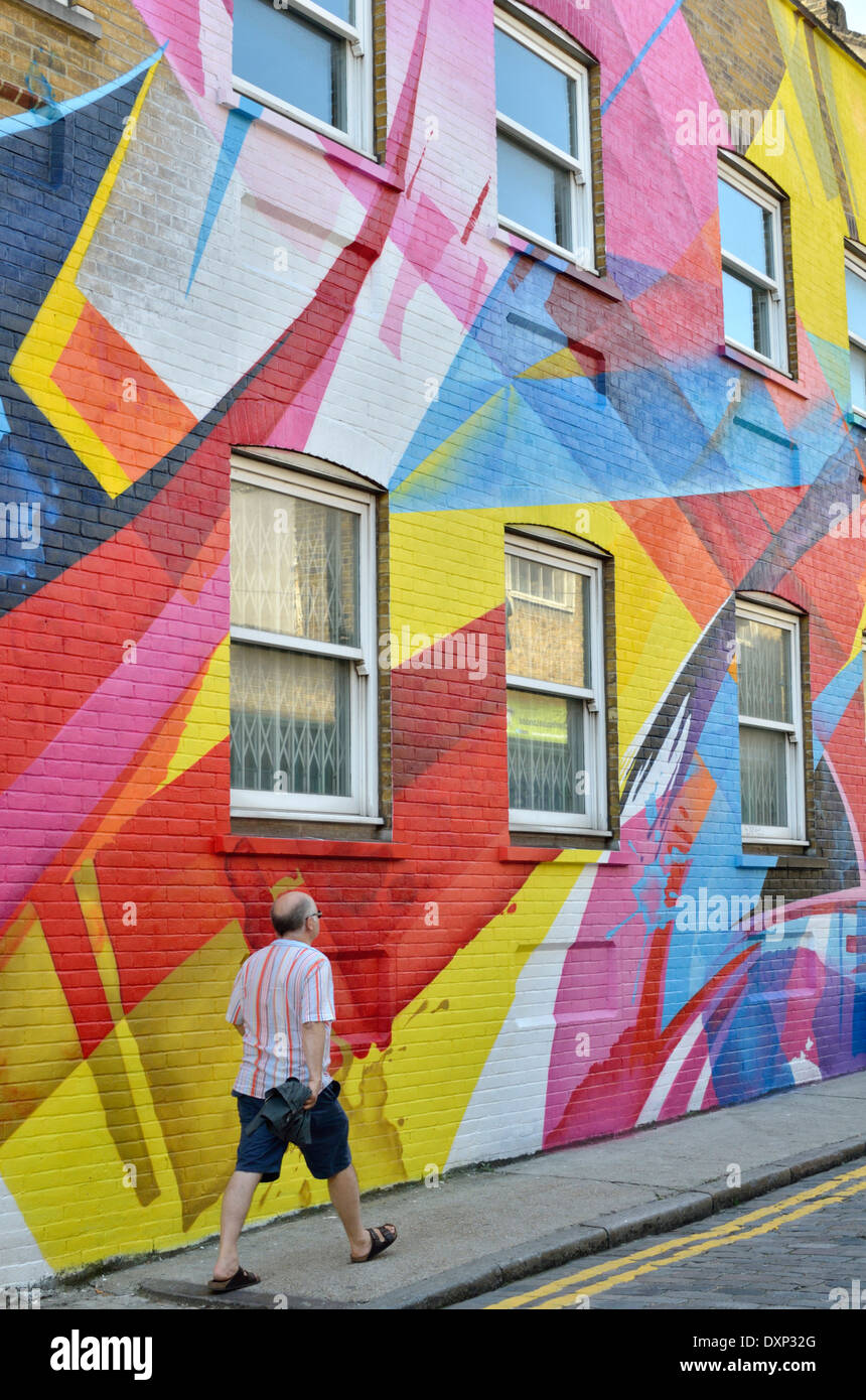 Un uomo a piedi passato un colorato astratto di pittura murale che copre il lato di un edificio, Shoreditch, Londra, Regno Unito. Foto Stock