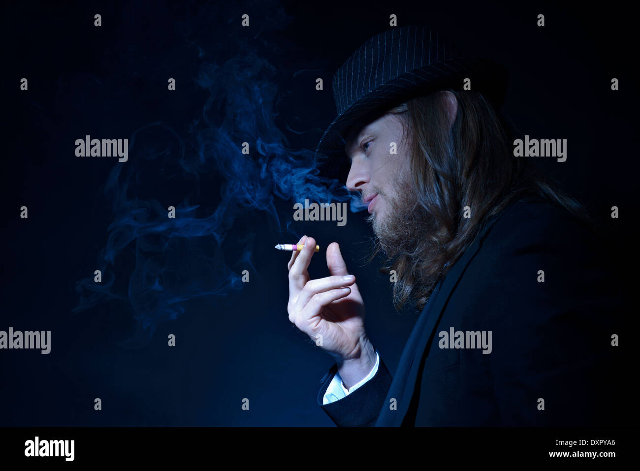 Un barbuto lunghi capelli uomo che indossa un cappello Fedora e tuta fumatori, un profilo laterale verticale con il sentiero di fumo, di un concetto creativo Foto Stock