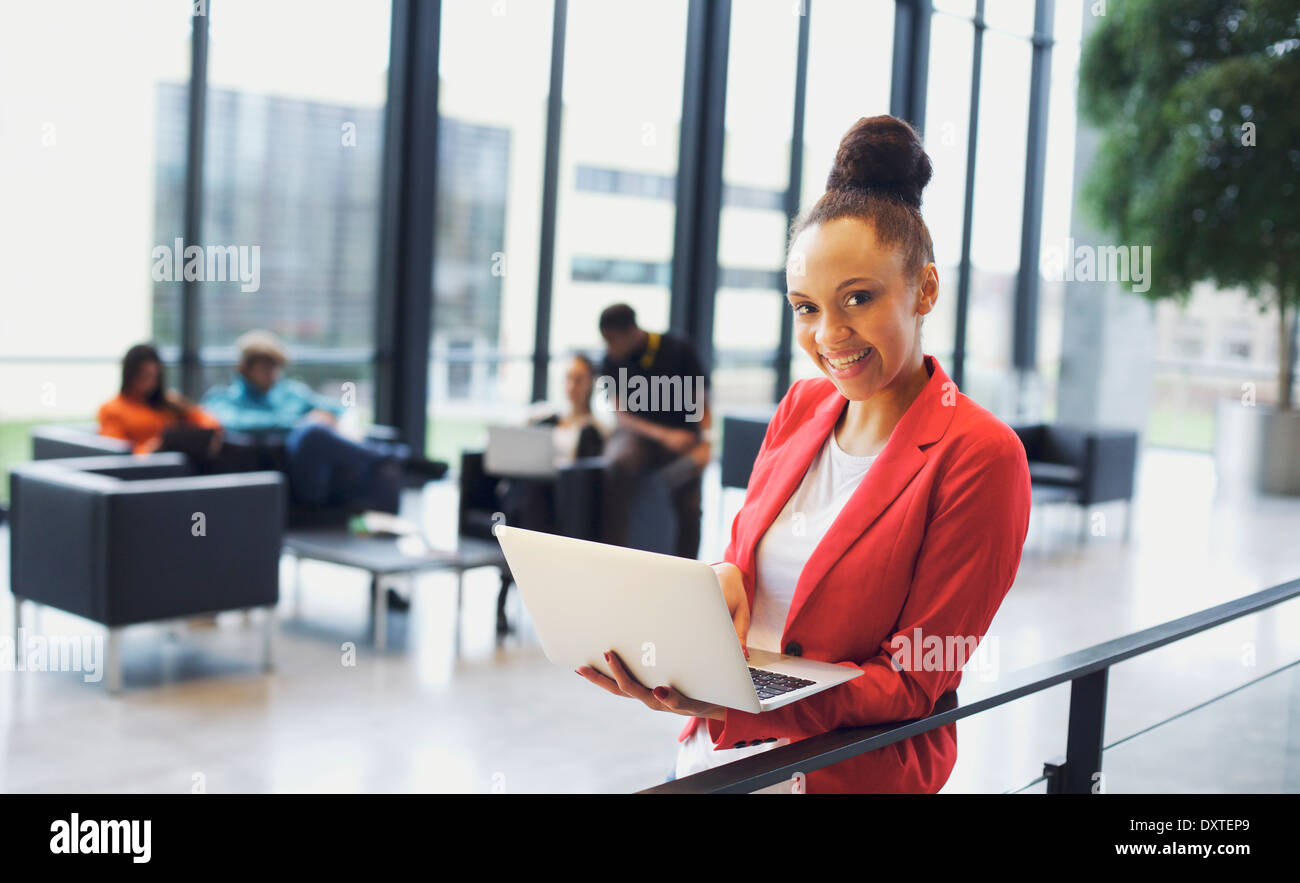 Giovane e bella donna con un computer portatile in ufficio moderno. African American imprenditrice in piedi da una ringhiera con persone che lavorano. Foto Stock