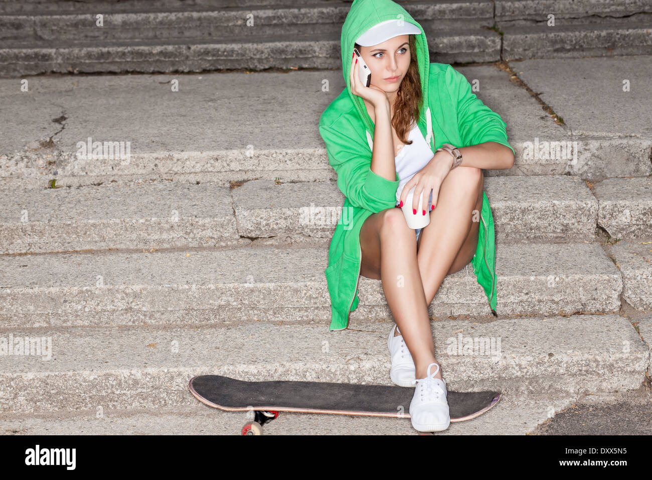 Giovane e bella ragazza in felpa con cappuccio verde colloqui sul cellulare seduti sulle scale di pietra con le sue gambe su skateboard Foto Stock