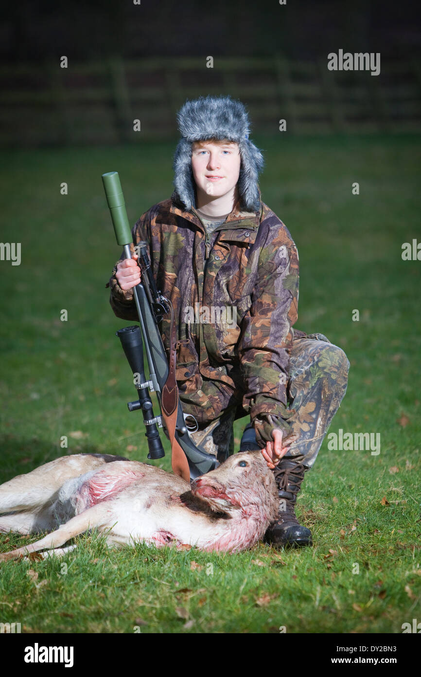 Un giovane cervo maschio stalker con un cervo morto e un fucile in un campo di erba al crepuscolo Foto Stock