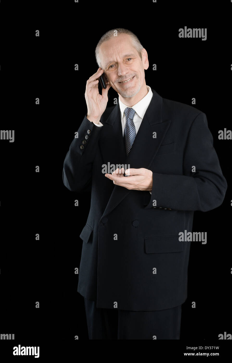 Un gradevole imprenditore indossando un abito nero di sorridere mentre si parla al telefono cellulare, su sfondo nero Foto Stock