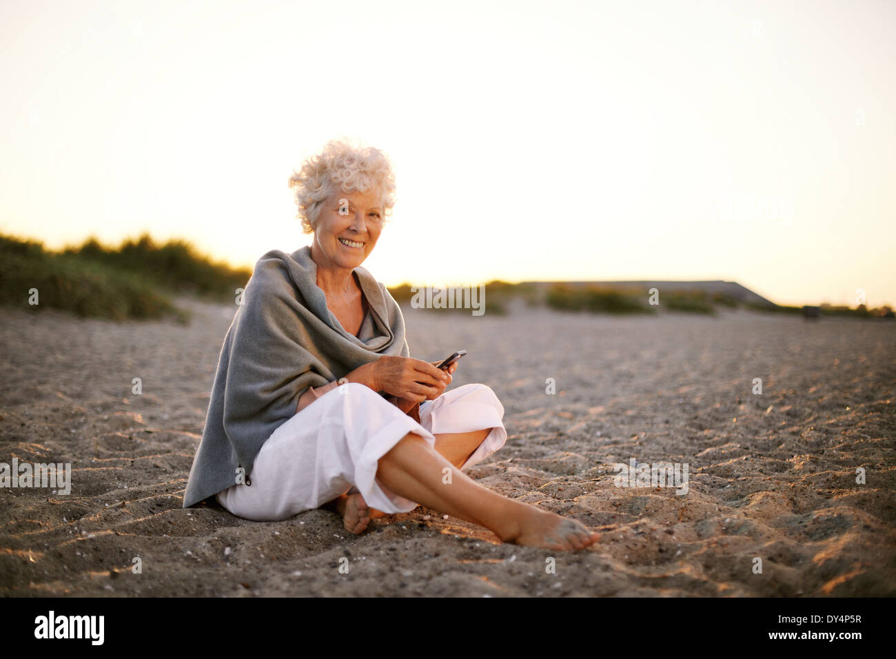 Felice pensionato donna seduta rilassante sulla spiaggia con un cellulare in mano. Senior donna caucasica con un telefono cellulare sulla spiaggia Foto Stock