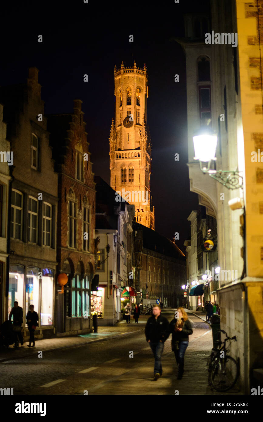 Belfry Tower illuminata di notte a Bruges Belgio // BRUGES, Belgio - Una strada medievale nel centro storico di Bruges conduce verso la torre illuminata del Belfry, un sito patrimonio dell'umanità dell'UNESCO. Il Belfry, costruito per la prima volta nel 1240 e completato nella sua forma attuale alla fine del XV secolo, sorge a 83 metri (272 piedi) sopra la piazza Markt. La suggestiva illuminazione notturna della torre mette in risalto i suoi dettagli architettonici gotici e sottolinea il suo ruolo di principale punto di riferimento della città. Foto Stock