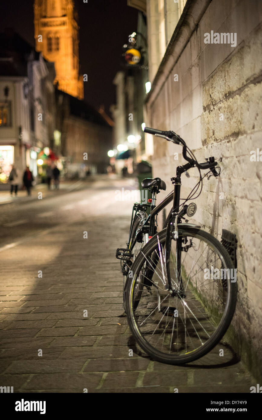 Bicicletta contro Wall Bruges Belgio di notte // BRUGES, Belgio — Una bicicletta solitaria si appoggia contro un edificio storico nel centro medievale di Bruges di notte. La scena cattura la miscela di trasporto moderno e architettura conservata caratteristica di questa città patrimonio dell'umanità dell'UNESCO. L'illuminazione stradale tradizionale crea un'atmosfera serale suggestiva lungo il paesaggio storico. Foto Stock