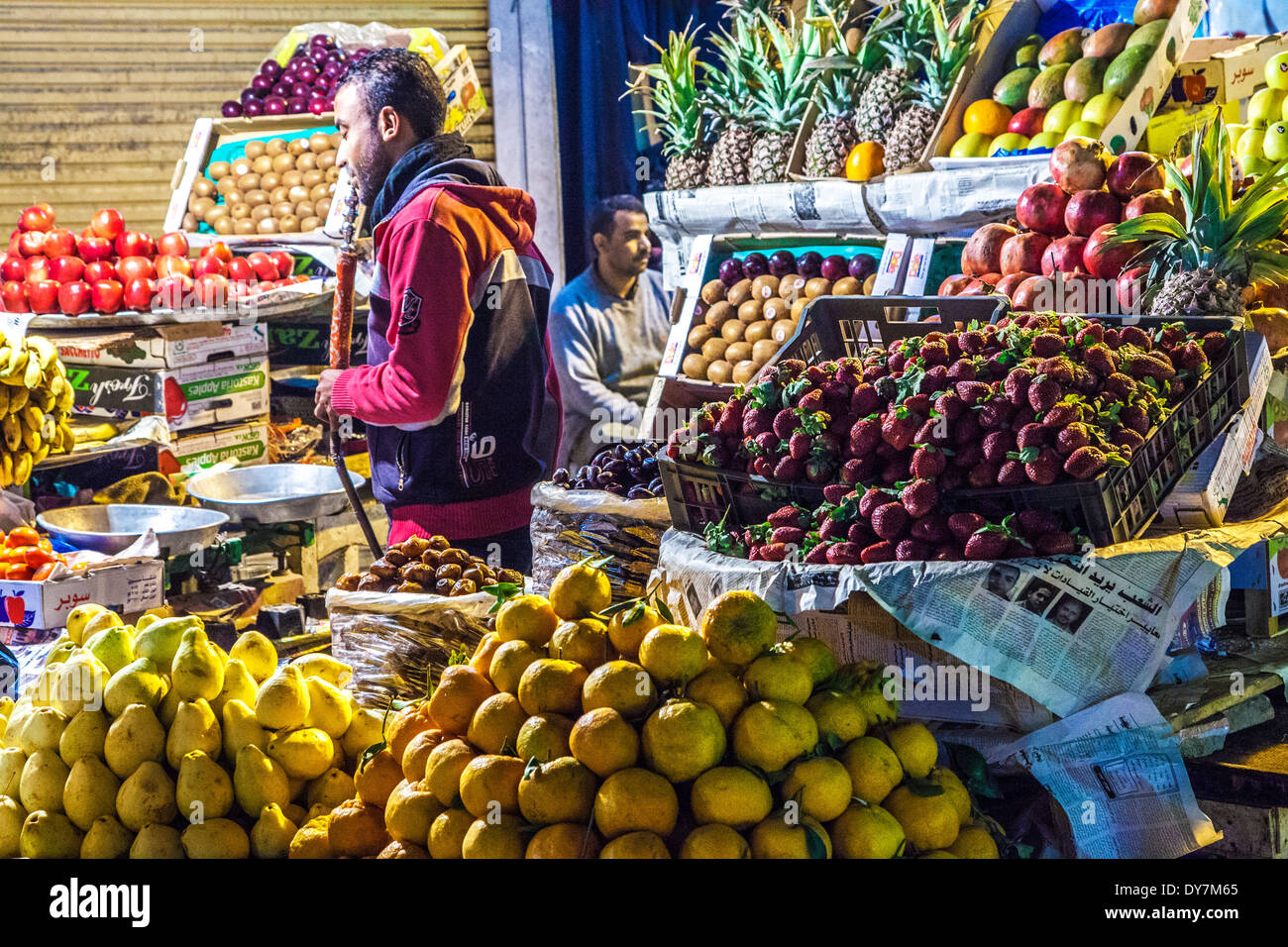 Stallkeeper fumare un narghilè dietro un frutto in stallo il mercato notturno o souk di Luxor, Egitto Foto Stock