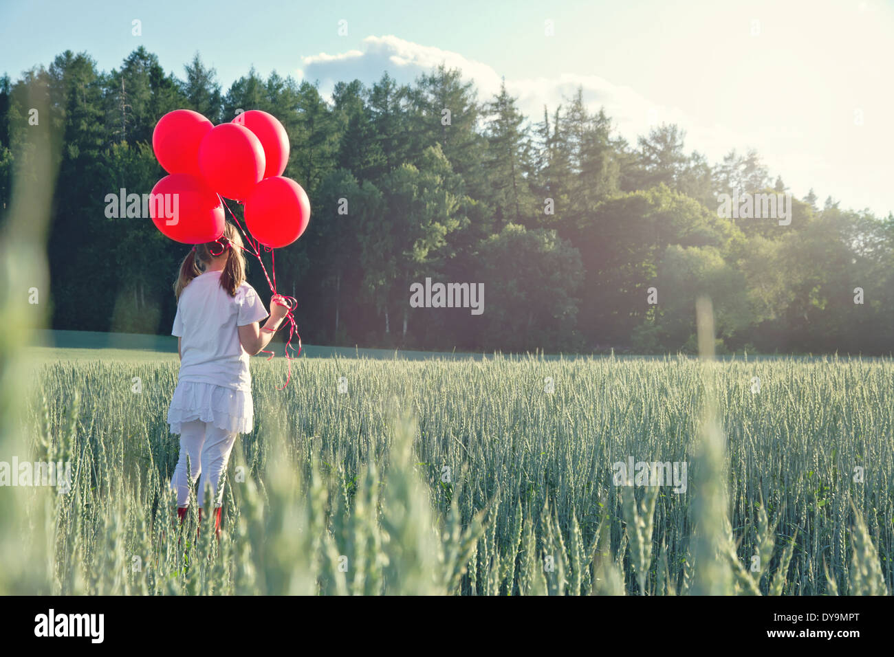 Bambina con un mucchio di palloncini rossi guardando il tramonto in un verde paesaggio estivo con nebuloso editing vintage Foto Stock
