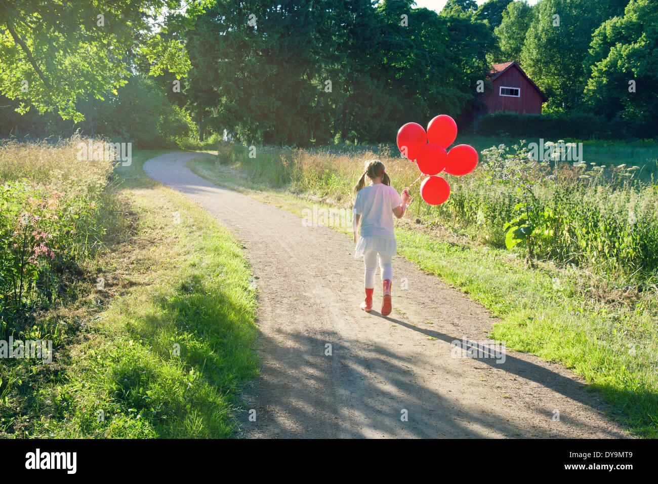 Bambina con un mucchio di palloncini rossi a camminare su una strada di campagna in un verde paesaggio estivo con nebuloso editing vintage Foto Stock