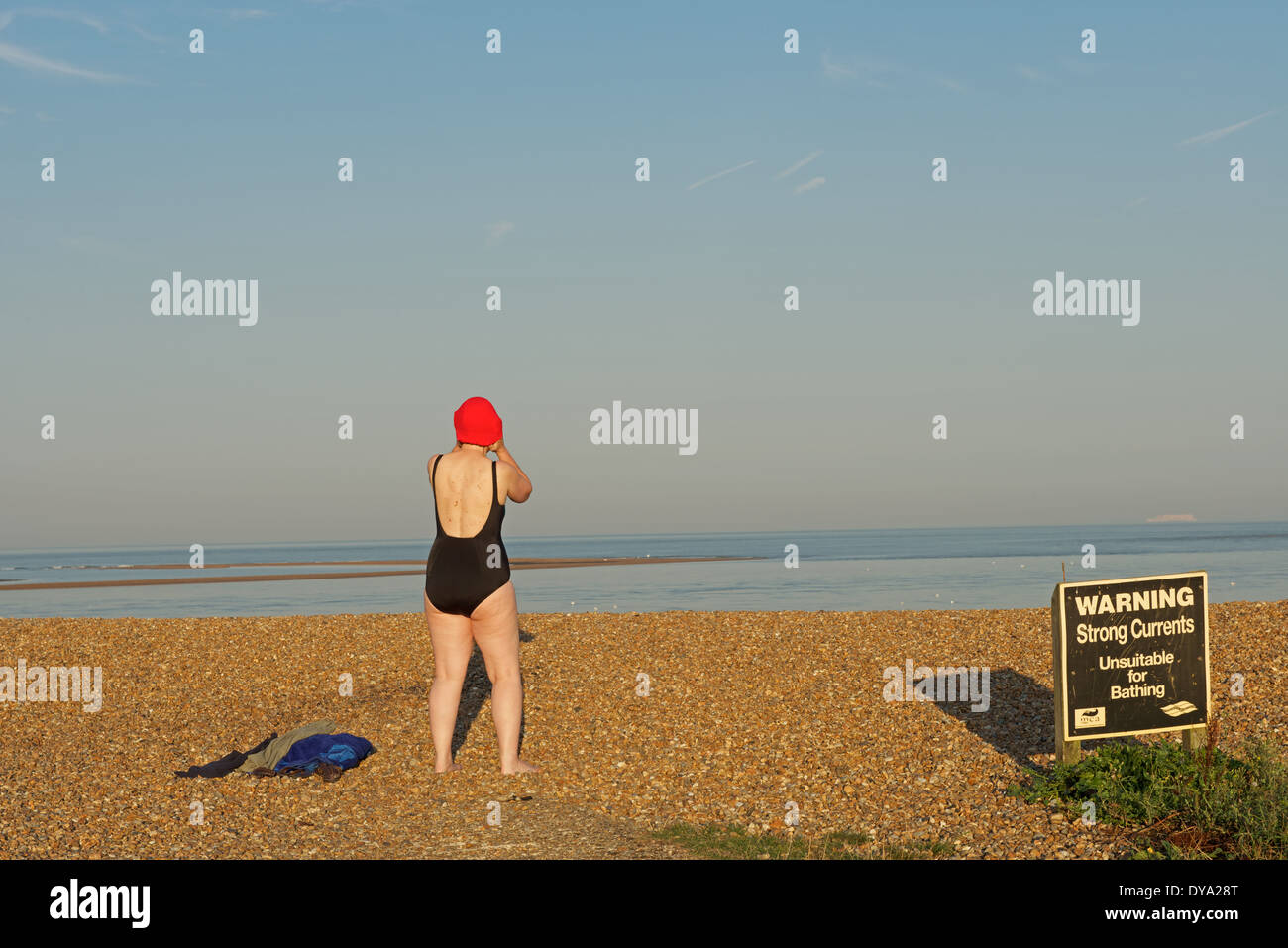 Donna preparando per una nuotata con un segnale di avvertimento di forti correnti e inadatto per la balneazione, strada di ciottoli, Suffolk, Regno Unito. Foto Stock