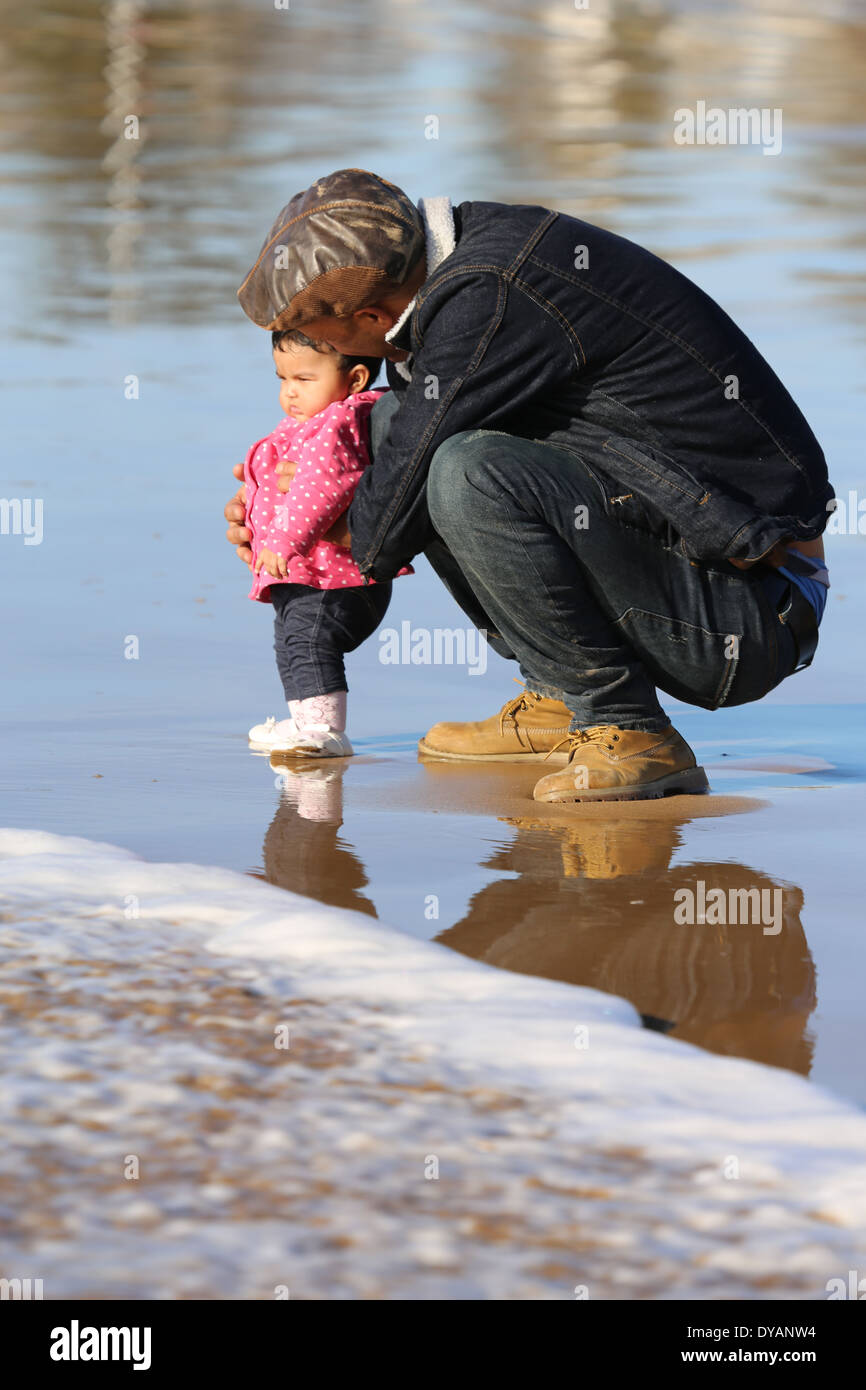 Padre coying oltre il toddler figlia su un wet spiaggia sabbiosa a Essaouira, Marocco, non vedendo un'onda in arrivo fino a molto ultimo minu Foto Stock