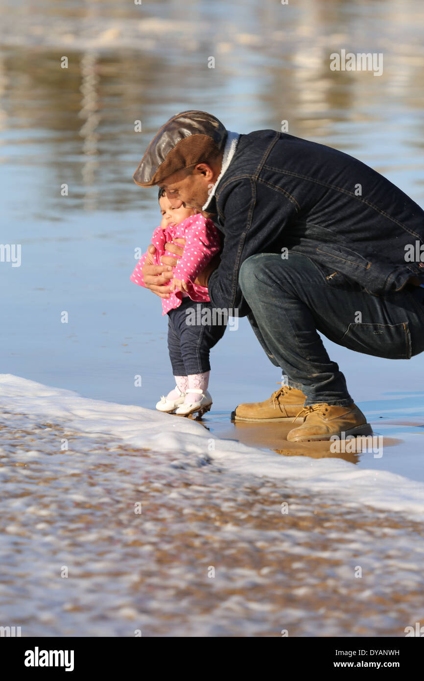 Padre coying oltre il toddler figlia su un wet spiaggia sabbiosa a Essaouira, Marocco, non vedendo un'onda in arrivo fino a molto ultimo minu Foto Stock