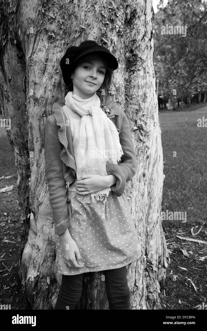 Una fotografia in bianco e nero di una giovane ragazza appoggiata contro un albero, indossando un cappello, abito, cardigan e sciarpa. Foto Stock