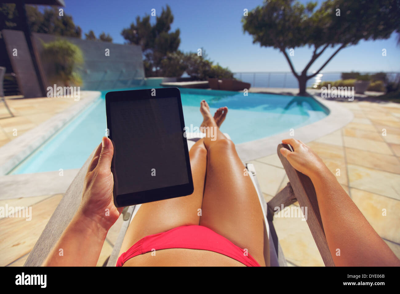 Giovane donna relax su una sedia a sdraio utilizzando un tablet PC vicino alla piscina. Utente POV. Modello femminile seduto su una sedia a sdraio. Foto Stock