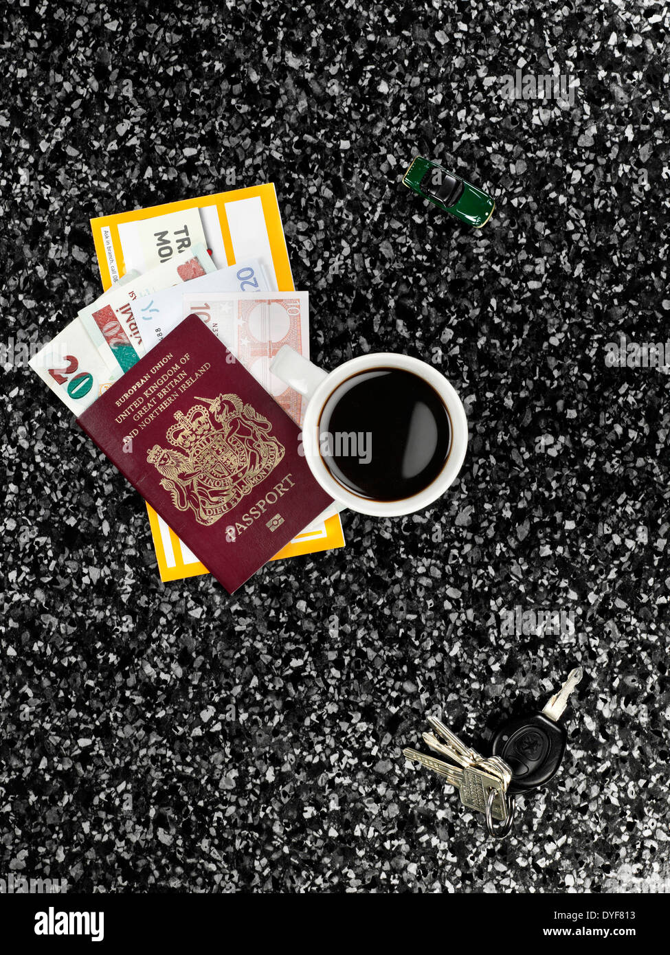 Un Ariel vista di alcuni tasti, tazza di caffè, GIOCATTOLO AUTO, passaporto e Denaro di viaggio al buio su un piano di lavoro in marmo Foto Stock