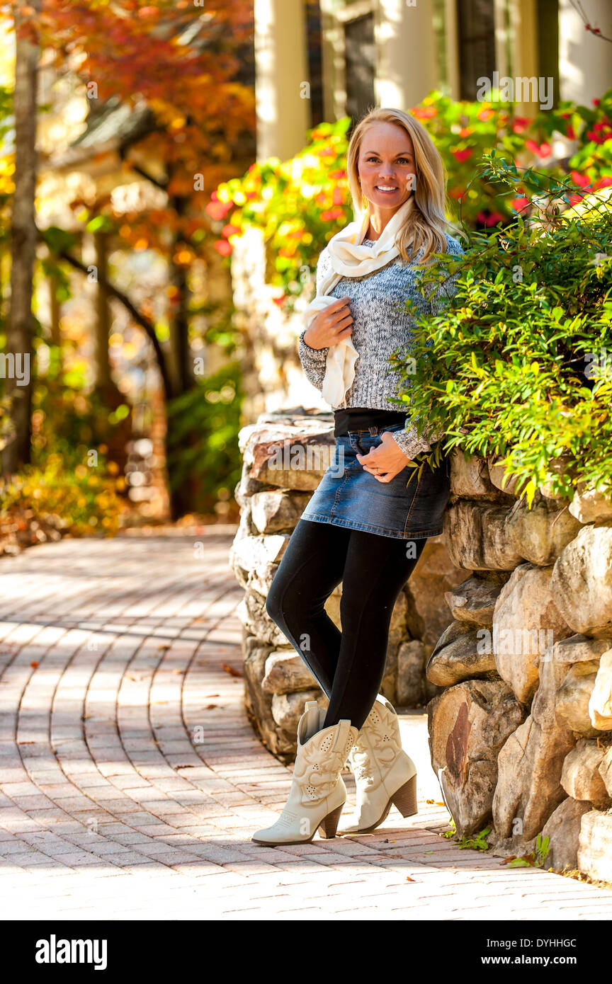 39 anni di donna bionda che indossa un maglione, mini gonna e stivali appoggiato su di un muro di pietra da un marciapiede in una strada suburbana in th Foto Stock